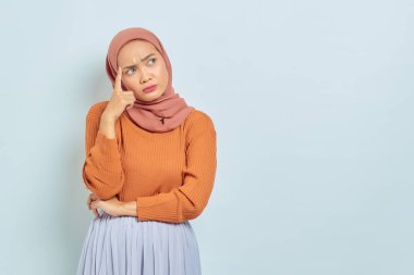 Kahverengi kazaklı, Asyalı genç bir Müslüman kadının portresi. Soruyu ciddi ciddi düşünüyor ve beyaz arka planda izole edilmiş fotokopi alanına bakıyor. Müslüman yaşam tarzı kavramı