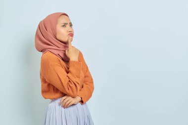 Kahverengi kazaklı ve tesettürlü düşünceli genç Asyalı Müslüman kadın beyaz arka planda izole edilmiş bir soruyu ciddi bir şekilde düşünüyor.