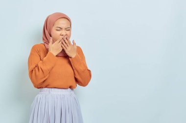 Kahverengi kazaklı ve tesettürlü güzel bir Asyalı kadın ağzını kapatıyor ve beyaz arka planda uykulu bir şekilde izole edilmiş hissediyor..