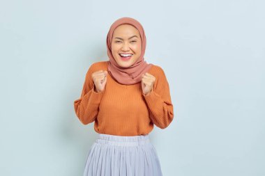 Kahverengi kazaklı, heyecanlı, Asyalı genç Müslüman bir kadın beyaz arka planda izole edilmiş bir şekilde evet diyerek zafer jesti yapıyor. Müslüman yaşam tarzı kavramı