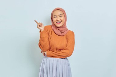 Kahverengi kazaklı, tesettürlü, tesettürlü, tesettürlü, reklam ürünlerini gösteren ve beyaz arka planda izole edilmiş kameraya bakan güzel bir Asyalı kadın..