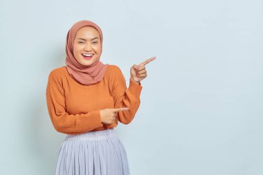 Kahverengi kazaklı, tesettürlü, tesettürlü, tesettürlü, reklam ürünlerini gösteren ve beyaz arka planda izole edilmiş kameraya bakan güzel bir Asyalı kadın..