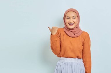Kahverengi kazaklı, tesettürlü, tesettürlü, tesettürlü, reklam ürünlerini gösteren ve beyaz arka planda izole edilmiş kameraya bakan güzel bir Asyalı kadın..