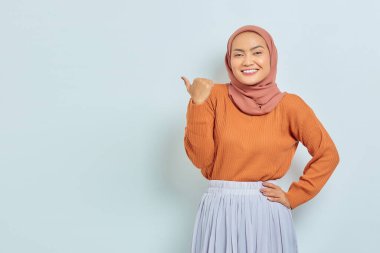 Kahverengi kazaklı, tesettürlü, tesettürlü, tesettürlü, reklam ürünlerini gösteren ve beyaz arka planda izole edilmiş kameraya bakan güzel bir Asyalı kadın..