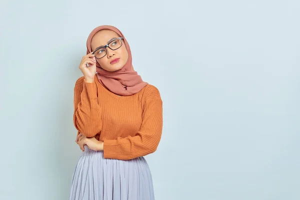 コピースペースを見て 白い背景に隔離された何かについて考えている茶色のセーターと眼鏡の若いアジアのイスラム教徒の女性 — ストック写真
