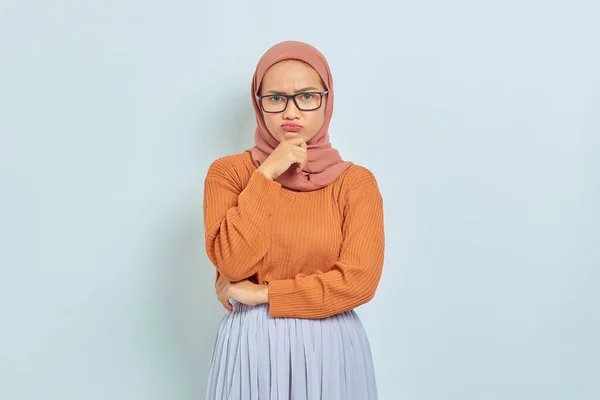 褐色のセーターと眼鏡を着たアジア系ムスリムの若い女性は 白い背景に孤立した何かを考えながら顎を保持します 宗教的なライフスタイルの概念は — ストック写真