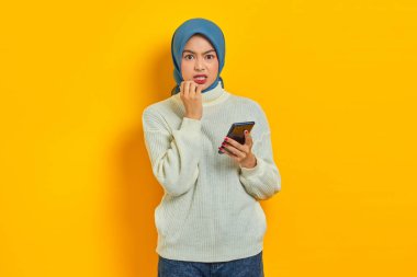 Endişeli, beyaz kazaklı ve tesettürlü genç Asyalı kadın cep telefonu kullanıyor ve sarı arka planda izole edilmiş ciddi bir ifadeyle kameraya bakıyor.