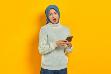 Beyaz kazaklı ve tesettürlü genç Asyalı kadın cep telefonu kullanıyor ve sarı arka planda izole edilmiş ağzı açık kameraya bakıyor. İnsanlar İslam dini kavramları