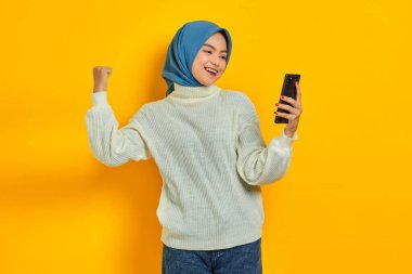 Beyaz süveter ve tesettürlü gülümseyen genç Asyalı kadın cep telefonu kullanıyor, sarı arka planda izole edilmiş kazanan hareketi yapıyor. İnsanlar İslam dini kavramları