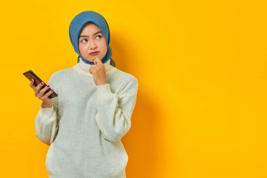 Beyaz kazaklı ve tesettürlü kafası karışık genç Asyalı kadının portresi sarı arka planda cep telefonunu tutarken çenesi üzerinde soru sormayı düşünüyor. İnsanlar İslam dini kavramları