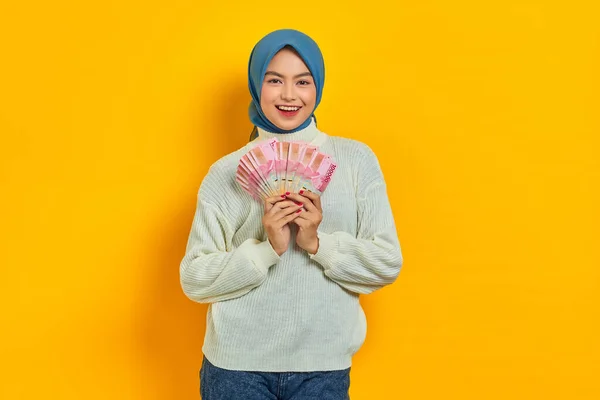 黄色の背景に孤立ルピア紙幣で現金のファンを保持白いセーターの陽気な美しいアジアのイスラム教徒の女性 人々の宗教生活の概念 — ストック写真