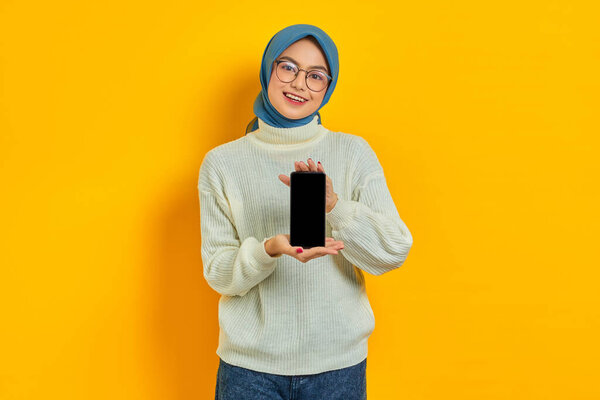 Smiling beautiful Asian woman in white sweater and hijab, showing blank mobile phone screen isolated on yellow background. Muslim lifestyle concept