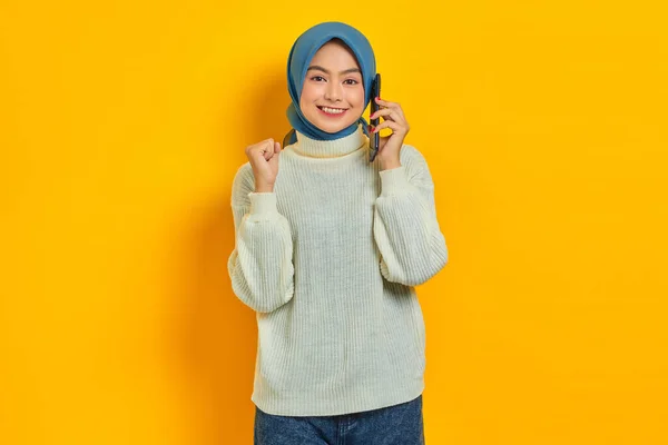 黄色の背景に隔離された幸運を喜びながら白いセーターを着た陽気な美しいアジアの女性が携帯電話を話しています 人々は宗教的概念を — ストック写真