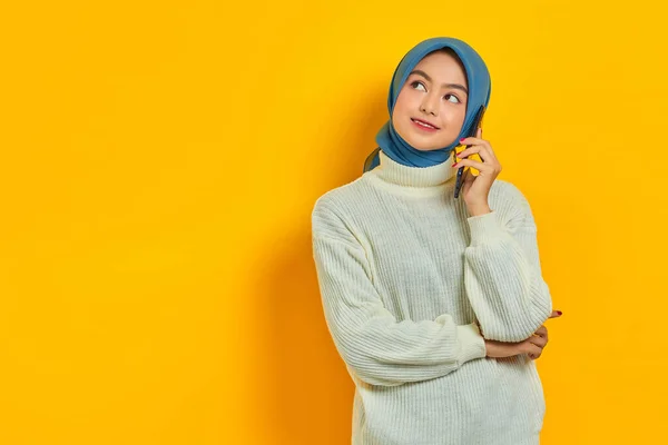 黄色の背景に隔離されたコピースペースを見ながら 白いセーターとヒジャーブを着た若いアジアの女性がモバイルで話しています 人々は宗教的概念を — ストック写真