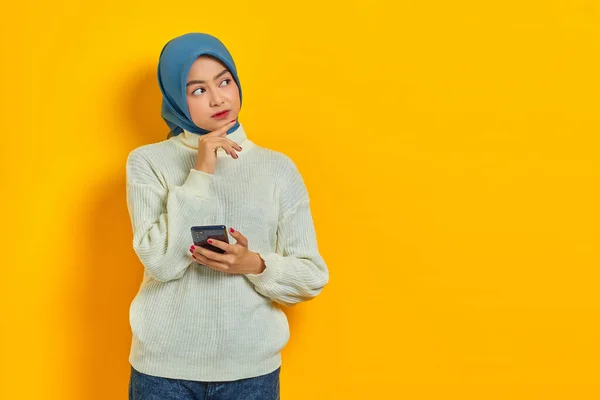 白いセーターを着た混乱した若いアジアの女性の肖像画と黄色の背景に隔離された携帯電話を保持しながら 顎に手で質問について考えてヒジャーブ 人々は宗教的概念を — ストック写真