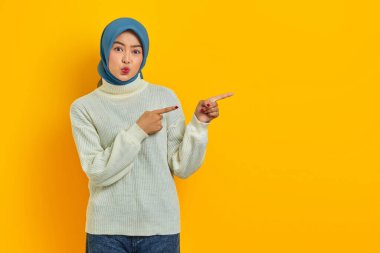 Beyaz kazaklı, dalgın Asyalı kadın ciddi ciddi soru sormayı düşünüyor ve sarı arka planda izole edilmiş parmakları işaret ediyor.