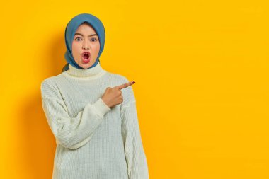Şok olmuş beyaz kazaklı güzel Asyalı kadın sarı arka planda izole edilmiş fotokopi alanını işaret ediyor.
