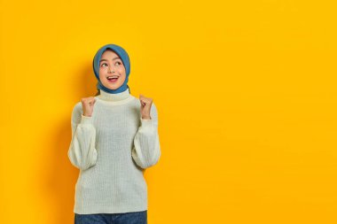 Heyecanlı beyaz kazaklı güzel Asyalı kadın sarı arka planda izole edilmiş yumruklarla büyük şans ve başarıyı kutluyor.