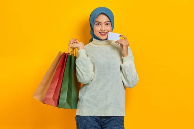 Beyaz kazaklı, neşeli Asyalı kadın elinde alışveriş torbaları ve sarı arka planda izole edilmiş kredi kartı tutuyor.