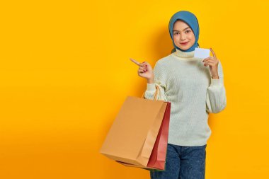 Beyaz kazaklı, elinde alışveriş çantası ve kredi kartı olan neşeli, güzel Asyalı kadın sarı arka planda izole edilmiş bir yeri işaret ediyor.