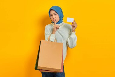 Beyaz kazaklı, elinde alışveriş çantası ve kredi kartı olan güzel Asyalı kadın sarı arka planda izole edilmiş sessizlik hareketi yapıyor.