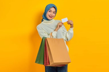 Beyaz kazaklı, neşeli Asyalı kadın elinde alışveriş torbaları ve sarı arka planda izole edilmiş kredi kartı gösteriyor.