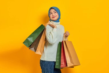 Beyaz kazaklı neşeli, güzel Asyalı kadın bir sürü alışveriş çantası gösteriyor. Sarı arka planda izole edilmiş indirimli reklam.