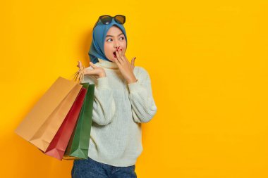 Şok olmuş beyaz kazaklı güzel bir Asyalı kadın, sarı arka planda elleri ağızlarında alışveriş torbaları tutuyor.