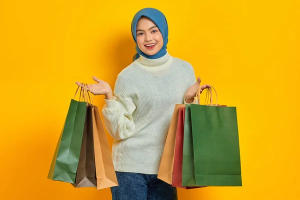 Cheerful beautiful Asian woman in white sweater showing so many shopping bags, shopping discount promo isolated over yellow background