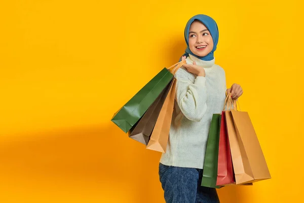 Cheerful beautiful Asian woman in white sweater showing so many shopping bags, shopping discount promo isolated over yellow background