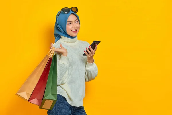 黄色の背景に隔離されたカメラを見て 携帯電話を使用して ショッピングバッグを保持白いセーターで陽気な美しいアジアの女性 — ストック写真
