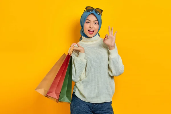 陽気美しいですアジアの女性でホワイトセーター保持ショッピングバッグ ショー大丈夫なジェスチャー絶縁上黄色の背景 — ストック写真
