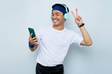Mavi bantlı ve beyaz tişörtlü gülümseyen Asyalı genç sporcu kulaklıklı favori müzik dinlerken, cep telefonuyla selfie çekerken, beyaz arka planda izole edilmiş barış jesti