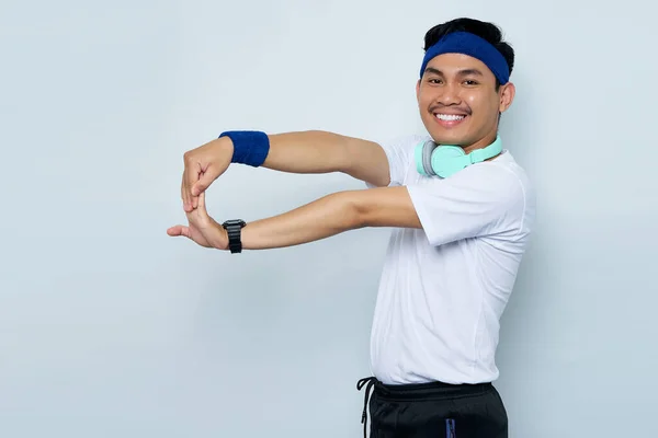 young Asian sportman in blue headband and sportswear white t-shirt with headphones, working out doing stretching exercising looking at camera isolated on white background. Workout sport concept