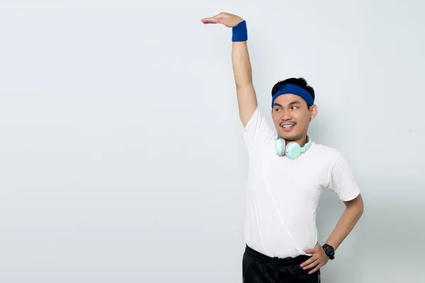 Portrait of smiling young Asian sportman in blue headband and sportswear white t-shirt with headphones, showing height measure isolated on white background. Workout sport concept