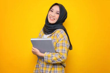 Smiling Young Asian Muslim student holding book, looking at camera isolated on yellow background. Back to school in high school college concept
