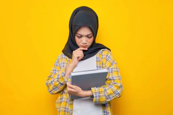 Pensive Young Asian Muslim student holding book, looks seriously thinking about a question  isolated on yellow background. Back to school in high school college concept