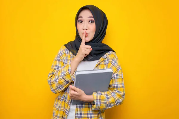 Smiling Young Asian Muslim student holding book showing silence gesture with finger isolated on yellow background. Back to school in high school college concept