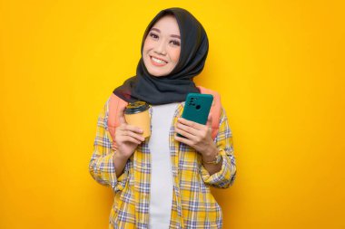 Sırtında cep telefonu, kahve ya da sarı arka planda izole edilmiş çay bulunan gülümseyen Asyalı Müslüman kadın öğrenci. Eğitim okulu üniversite konsepti