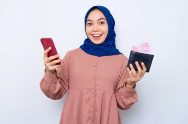 Pembe gömlekli, cep telefonu tutan ve beyaz arka planda izole edilmiş cüzdan gösteren neşeli Asyalı genç Müslüman kadın. İnsanlar dini yaşam tarzı konsepti