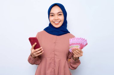 Pembe gömlekli gülümseyen Asyalı müslüman kadın cep telefonu kullanıyor ve elinde beyaz arka planda izole edilmiş para banknotları tutuyor. İnsanlar dini yaşam tarzı konsepti