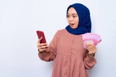 Pembe gömlekli genç Asyalı Müslüman kadın cep telefonu kullanıyor ve elinde beyaz arka planda izole edilmiş para banknotları tutuyor. İnsanlar dini yaşam tarzı konsepti