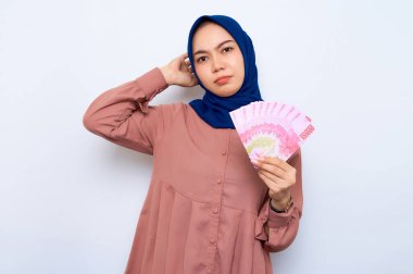 Kafası karışmış pembe gömlekli, Asyalı Müslüman bir kadın elinde beyaz arka planda izole edilmiş para banknotları tutuyor. İnsanlar dini yaşam tarzı konsepti