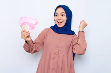 Heyecanlı pembe gömlekli, Asyalı genç Müslüman kadın beyaz arka planda izole edilmiş para banknotları tutuyor. İnsanlar dini yaşam tarzı konsepti
