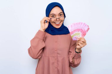Heyecanlı pembe gömlekli, Asyalı genç Müslüman kadın beyaz arka planda izole edilmiş para banknotları tutuyor. İnsanlar dini yaşam tarzı konsepti