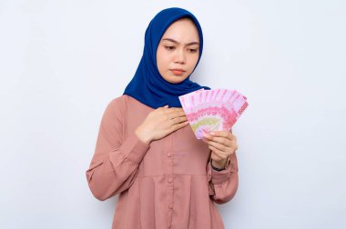 Kafası karışmış pembe gömlekli, Asyalı Müslüman bir kadın elinde beyaz arka planda izole edilmiş para banknotları tutuyor. İnsanlar dini yaşam tarzı konsepti