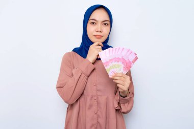 Kafası karışmış pembe gömlekli, Asyalı Müslüman bir kadın elinde beyaz arka planda izole edilmiş para banknotları tutuyor. İnsanlar dini yaşam tarzı konsepti