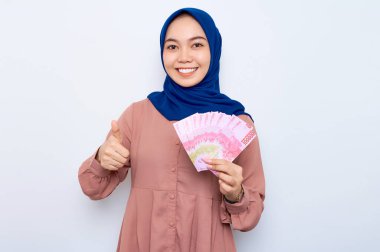 Pembe gömlekli gülümseyen Asyalı müslüman kadın elinde para banknotları ve beyaz arka planda soyutlanmış baş parmağını gösteriyor. İnsanlar dini yaşam tarzı konsepti