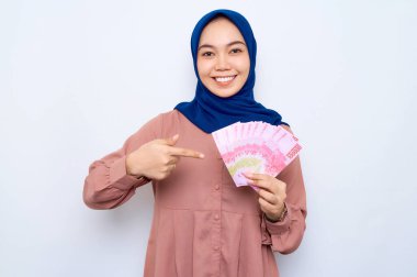 Heyecanlı, pembe gömlekli, Asyalı genç Müslüman kadın beyaz arka planda izole edilmiş para banknotları gösteriyor. İnsanlar dini yaşam tarzı konsepti