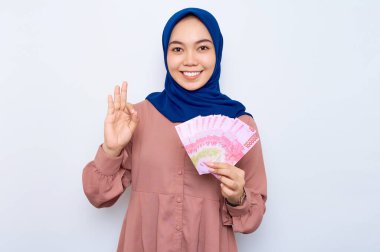 Pembe gömlekli gülümseyen Asyalı müslüman kadın elinde para banknotları ve beyaz arka planda izole edilmiş işaret işareti tutuyor. İnsanlar dini yaşam tarzı konsepti